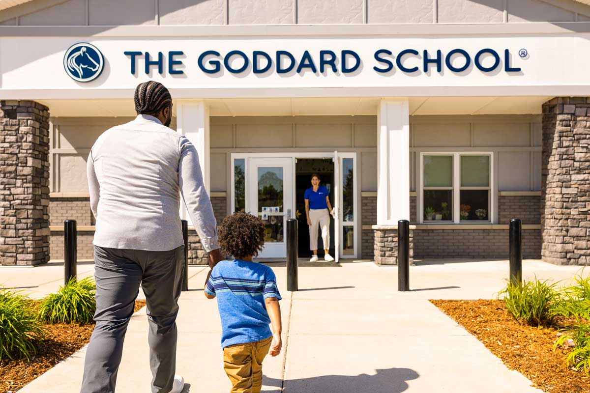 A father and son holding hands walking up to the front door of a Goddard School where a teacher is waiting to welcome them into the school