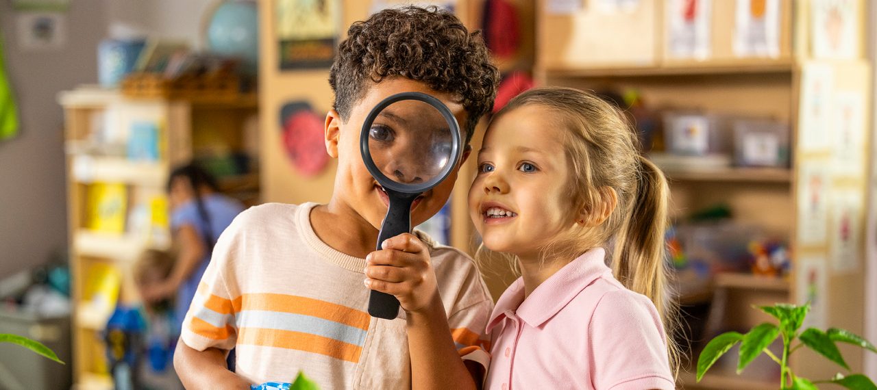 Two children looking through a magnifying glass in a preschool classroom